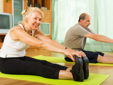 Two people are seeing reaching out their arms to touch their twos while sitting on the floor on exercise mats.