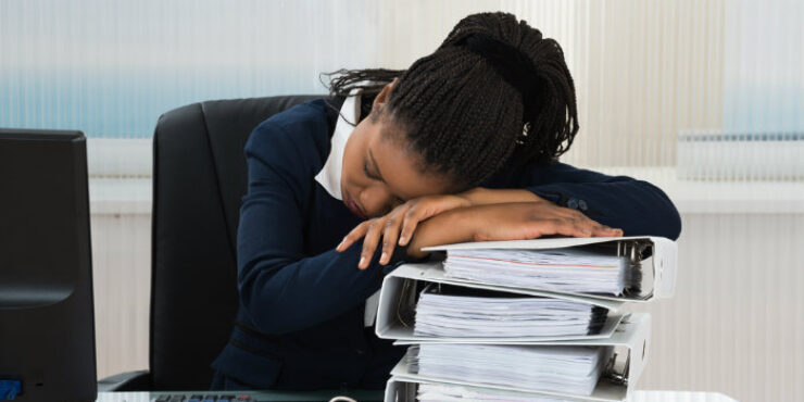 A woman rests her head on a stack of binders at a desk, appearing fatigued.