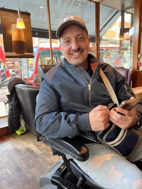 MS and walk | Multiple Sclerosis News Today | A closeup shows 43-year-old Chris Pamayides, who has RRMS, smiling and sitting in a scooter at a London coffee shop. He's holding his phone in his left hand.