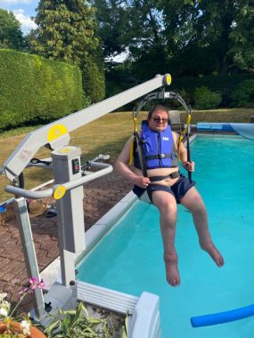 uk heat wave | Multiple Sclerosis News Today | John sits in a hoist chair hovering above a swimming pool. He's wearing a life jacket and sunglasses