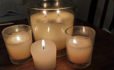 A close-up photo shows one large candle with two wicks and three smaller candles with one wick each. All five flames are lit, and the room is dark.