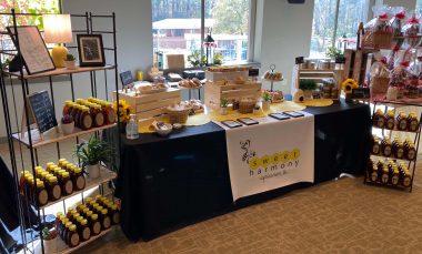 An indoor specialty market stand shows various artisanal products including honey, soaps, and other items. The predominant color on the products and banner is yellow