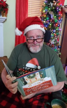 This photo shows a man in a Santa hat, holding the book "The Night Before Christmas" in his hands. A Christmas tree is behind him, and a poinsettia is visible on a mantel over his shoulder. He's sitting, has a blond, graying beard, and wears a green T-shirt with a Santa hat on it. 