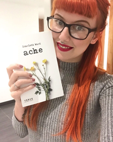 A young, red-haired, white woman who appears to be in her early 20s, and wearing big, black, designer glasses and red lipstick, smiles at the camera while holding up a book that partially says "ache." She is wearing a gray turtleneck sweater and is sitting on either a couch or a chair in a nondescript room. 