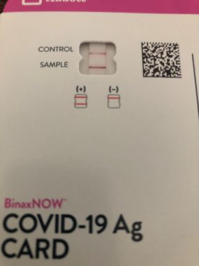 A close-up photo of a COVID-19 Ag Card shows two pink lines, indicating a positive test.