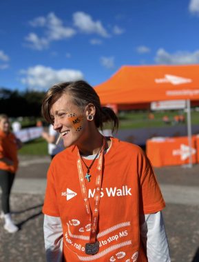 A woman wears an orange T-shirt for the MS Society's MS Walk over a long-sleeve white shirt. She has a medal with an orange ribbon around her neck and is wearing a cross necklace. Her hair is tied back in a low bun, and she has black and orange face paint on one cheek. She's looking to her right and laughing while standing outside. An orange canopy and a grassy field are visible behind her.
