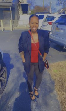 A Black woman in a black sport coat, red blouse, black jeans, and pretty shoes stands in an outdoor parking lot with a purse wrapped around her body and her hands in her blazer's pockets. The sun shines on her and she's wearing a broad smile. 