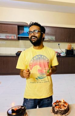A youngish band bearded Indian man in a yellow surfing T-shirt and wearing glasses flashes a wide, open-mouth smile as he looks to his right. He is standing in a kitchen in front of two cakes with candles on them, presumably to mark the anniversary of his MS diagnosis three years ago. He is holding a knife to cut the cakes in his left hand. 