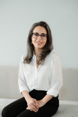 A professional portrait shot of a middle-aged white women wearing black glasses and sitting before a white background. She is wearing a white blouse and black dress pants, has her hands folded on her lap, and is wearing a broad smile