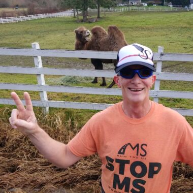A man poses in a field outside, with a white fence and a camel behind him. He is wearing an orange "Stop MS" shirt, a trucker hat, and dark sunglasses, and is making a peace sign with two fingers on his right hand.