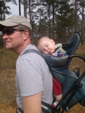 In the foreground of some open grass and a stand of pine trees, a seemingly fit man with short brown hair in a gray T-shirt, gray hat, and sunglasses is seen in profile, strapped on his shoulders into a blue back carry, where a baby in red leggings is sleeping. 
