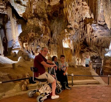The author sits on his electric mobility scooter next to his two grandchildren inside Luray Caverns. They're on a flat, paved brick surface, and behind them is a wall of shadowy stalactites and stalagmites.