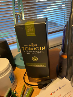 An unopened box with a bottle of Tomatin 12-year single malt Scotch inside sits on a desk cluttered with items. 