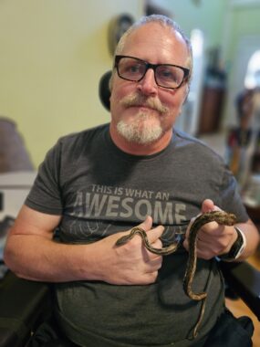 A middle-aged man is holding up a small, gray rat snake. He's wearing a gray graphic T-shirt and glasses, and is seated in his wheelchair in what appears to be his living room.