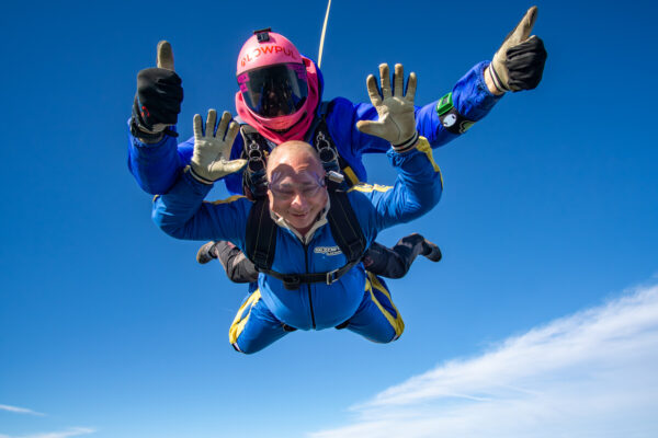 Two tandem skydivers dressed in blue skydiving suits with yellow stripes are in mid-jump in the sky. The instructor wears a pink helmet and flashes two thumbs up; the tandem diver has his arm extended above his head, no helmet, and wears a broad smile. 