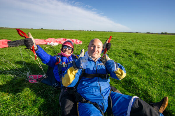 Two skydivers - one an instructor and one a tandem diver - sit on the ground in a vast, green field, both giving the thumbs up sign. A deployed parachute is spread out on the ground behind them. They are both smiling, having completed a successful jump from a plane.