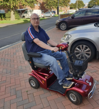 A bald, middle-aged man wearing a blue shirt, jeans, and sits on a red mobility scooter in a bricked parking lot, with two cars in the background. It looks to be next to a road in a residential neighborhood.