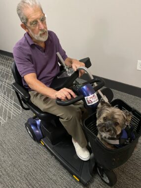 A man with gray hair, beard, mustache, and wire glasses drives a black scooter on a gray carpet. He wears a purple polo shirt, khakis, and white shoes. A small dog sits in the front basket of the scooter.