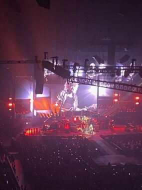 A photo taken from nosebleed seats shows the band Queen performing onstage. The venue is dark but the stage is lit up in red and purple lights. 