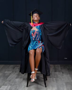A woman, sitting on a stool, poses in her cap and gown with her arms outstretched. 