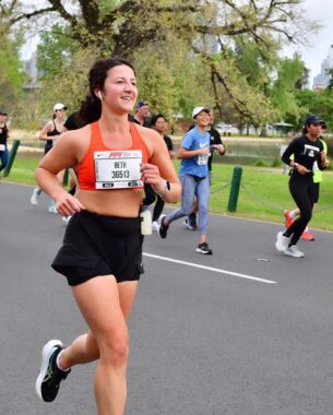 A woman in an orange sports bra and black shorts smiles while running down a road amid a group of other runners. She has a white tag attached to her front that displays her name, Beth, and marathon ID number. 
