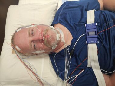 A man lies prone and strapped in to a hospital-type bed, with a series of wires connected to his head and oxygen tubes running to his nose. He looks up at the camera overhead and smiles. 