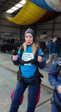 A woman, giving a thumbs up, stands near an open parachute, dressed in skydiving gear. 