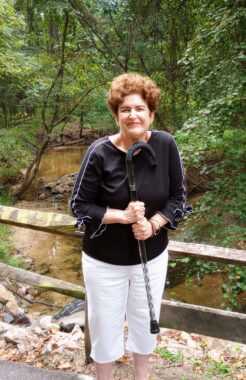 A woman poses on a nature trail and holds her cane up with both hands. 