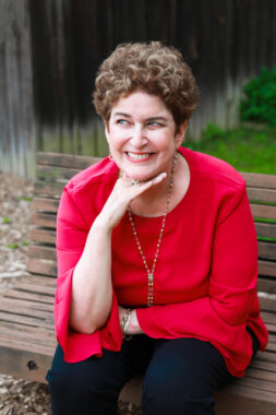 A woman with short curly hair poses for a photo while sitting on a park bench. She's wearing a bright-red shirt and smiling while looking off to the side.