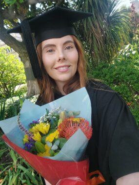 A woman, wearing a graduation cap, smiles as she holds flowers. 
