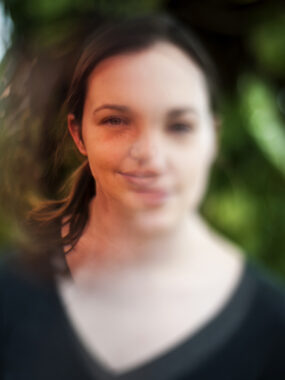 A headshot of a woman is edited to depict how she views the world. Only part of her face is in focus, and the coloring of that part appears more saturated.