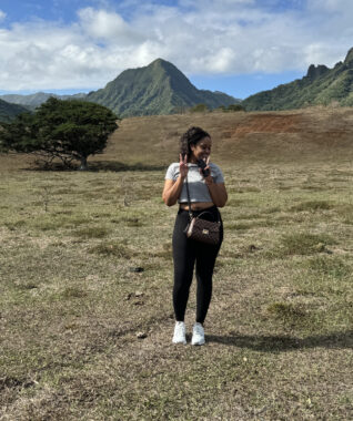 A woman stands in the middle of an expansive field, with mountains towering in the background. She's looking off to the side and making peace signs with both hands.