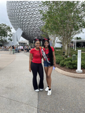 A mother and daughter pose for a photo at Disney World, wearing matching red shirts and Mickey Mouse ears. The daughter is wearing a birthday sash.
