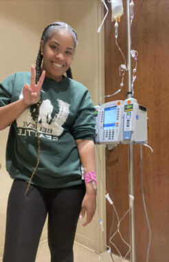 A woman stands next to an IV pole while receiving an infusion in her arm. She's smiling and making a peace sign with her right hand.