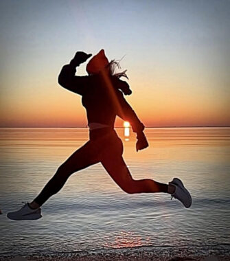 A woman jumps in the air with a sunrise over a body of water in the background.