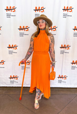 A woman with tattoos poses at a National MS Society event. She's wearing an orange dress and holding a matching orange purse and cane.