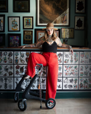 A woman poses against a counter in a tattoo shop. She's wearing a hat, a black top, and red pants, and one foot is propped up on a sleek rollator.