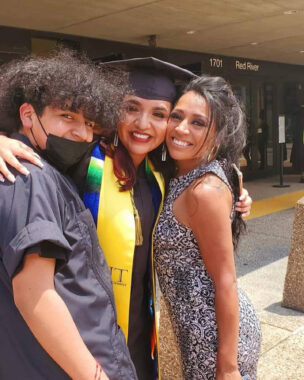A young woman wearing a black graduation cap and gown stands between her mom and her brother, with one arm around each of the. The mom is wearing a black-and-white dress and has her hair pulled back, while the brother is wearing a short-sleeve shirt and black face mask. All three are smiling.