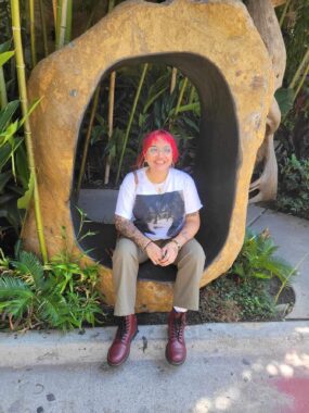 A young woman with pink hair and glasses sits on a sculpture outside the Dallas World Aquarium.