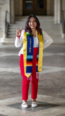 A young woman poses for a college graduation photo. She's wearing a gold sash from the University of Texas at Austin and a Latinx stole, and is making the longhorns sign with her right hand.