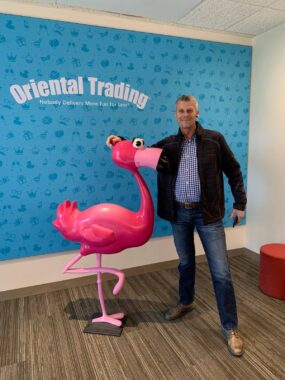 A man in a black jacket and casual dress clothes stands next to a large pink flamingo doll, in front of a blue wall that says "Oriental Trading." 