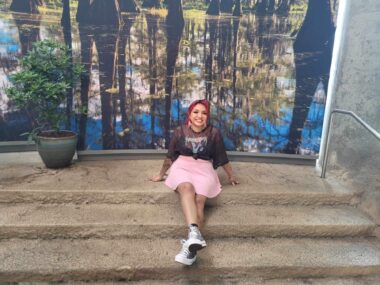 A young woman with pink hair sits on stone steps outside of an aquarium. She's wearing a black top and a pink skirt with her legs stretched out in front of her, and she's smiling broadly.