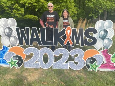 A father and his adult daughter stand next to each other outdoors, behind a giant sign that says "Walk MS 2023." The two are also wearing matching black T-shirts that say "Fight Multiple Sclerosis." Behind them is a row of trees. 