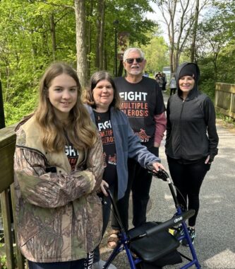 A multigenerational family of four that includes three women and one man, stand on an outdoor path with light jackets on. They're wearing black T-shirts that say "Fight Multiple Sclerosis." A row of trees lines the path. 