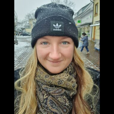 A close-up of a woman with long, blond hair, wearing an Adidas winter cap, a scarf, and a winter jacket. She is standing outdoors in a snow-covered city.