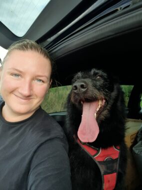 A close-up of a woman and her black dog inside a vehicle. The dog has a long tongue hanging out. 