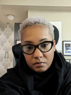 A woman with short bleached hair takes a photo at her desk.