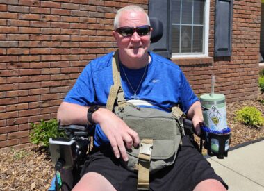 A middle-aged man smiles for a photo on a sidewalk in front of what appears to be a brick house. He's wearing a blue T-shirt, shorts, and dark sunglasses and is seated in his power wheelchair. A large crossbody bag is slung over his right shoulder and rests on his lap.