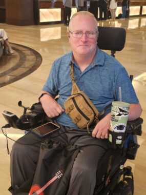 A middle-aged man is seated in his power wheelchair in what appears to be a large lobby with wooden floors. He's wearing a blue collared shirt and has a tan utility fanny pack slung over his right shoulder, so that the bag rests at the level of his abdomen. His phone is resting on his right thigh, and there's a large green tumbler in his wheelchair's cup holder to his left.