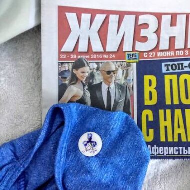 A close-up photo shows a white pin featuring an iris and the words "New Life." It's attached to a blue piece of fabric, perhaps a shirt or a sweater. Both are lying atop a June 2016 edition of the Russian newspaper "Zhizn," meaning "Life."